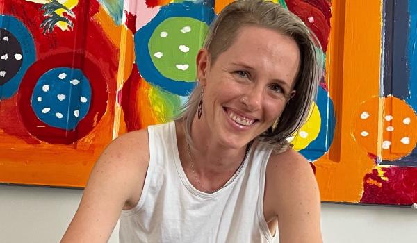 A blond white person with medium length hair with an undercut, a nose piercing, and wearing a white tank top. They are smiling.